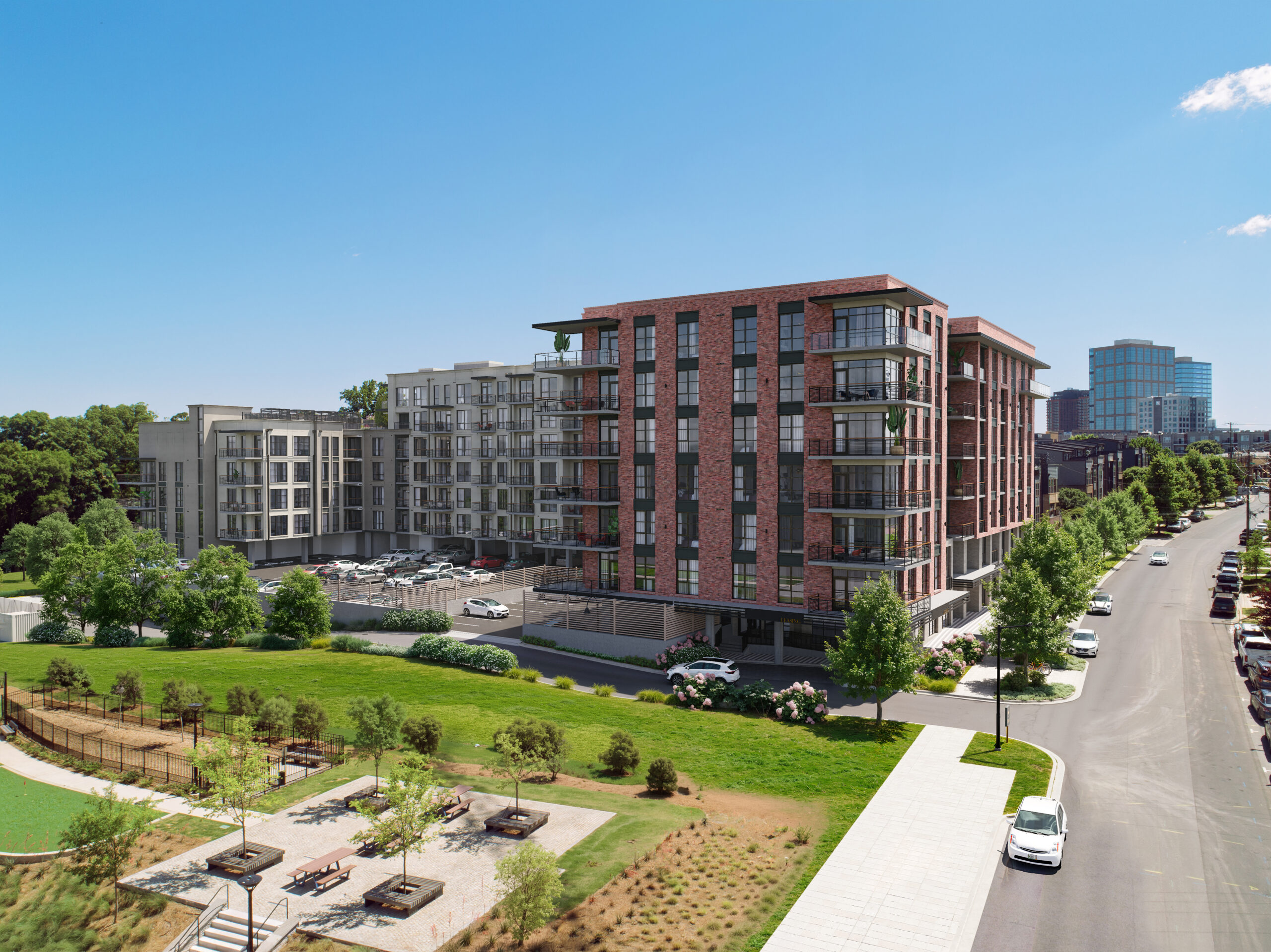 Modern apartment buildings with balconies overlook a green park and street lined with trees and parked cars on a sunny day.