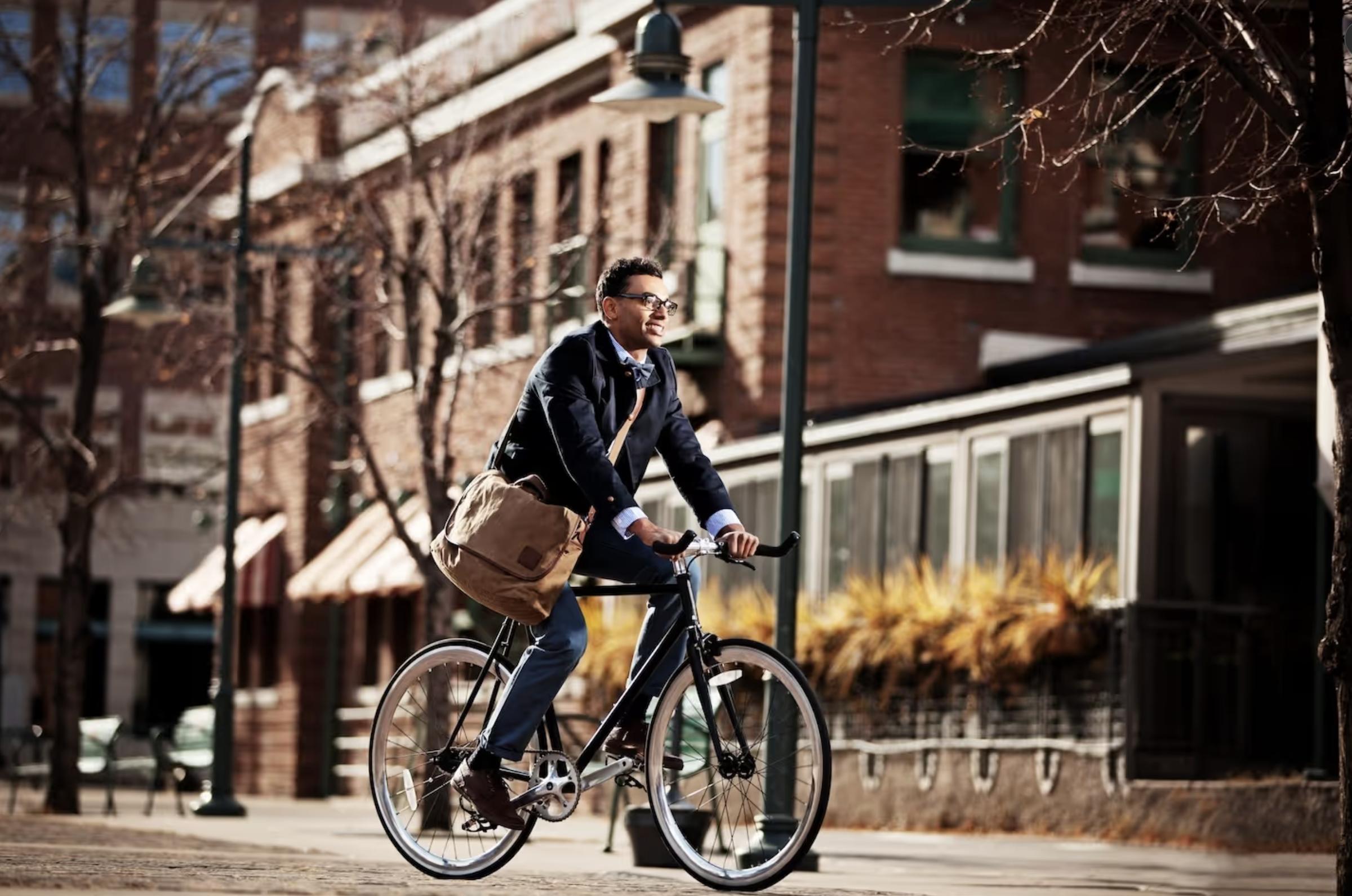 A man in business attire rides a bicycle with a messenger bag in an urban area on a sunny day.