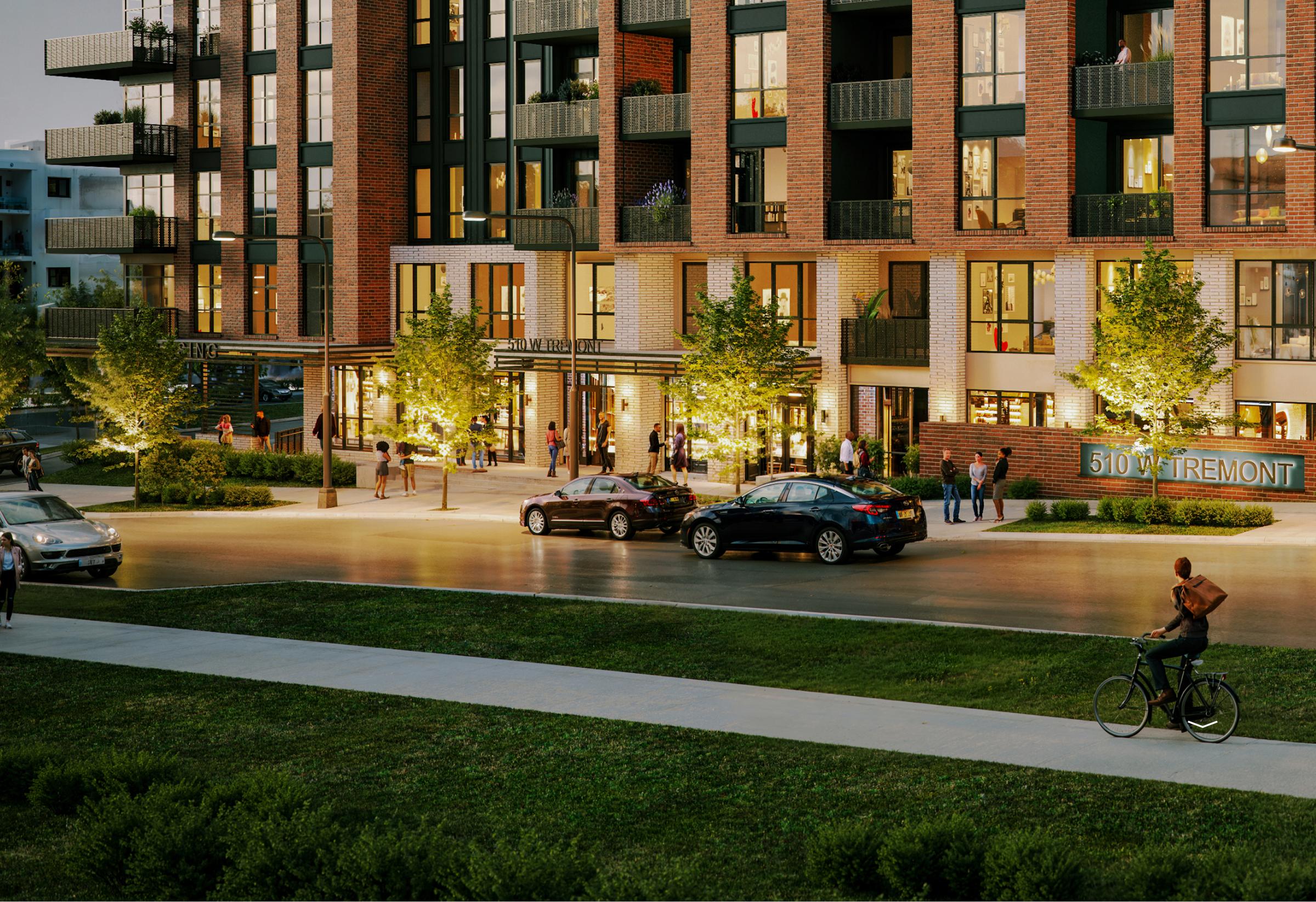 A cyclist rides past a modern apartment building at dusk, with people gathered outside and cars parked along the street.