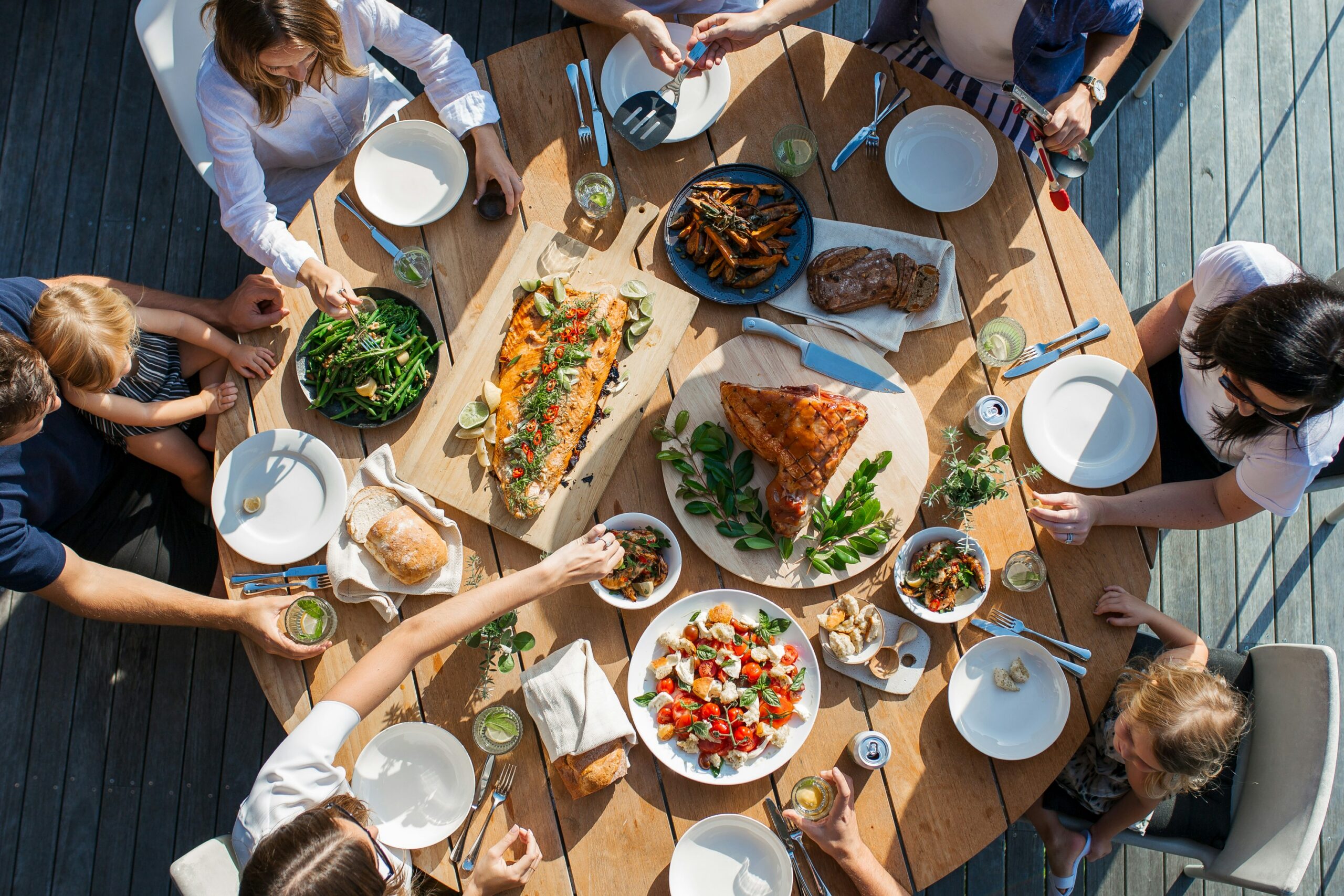 People gathered around a wooden table outdoors, sharing a meal with salads, bread, fish, and drinks.
