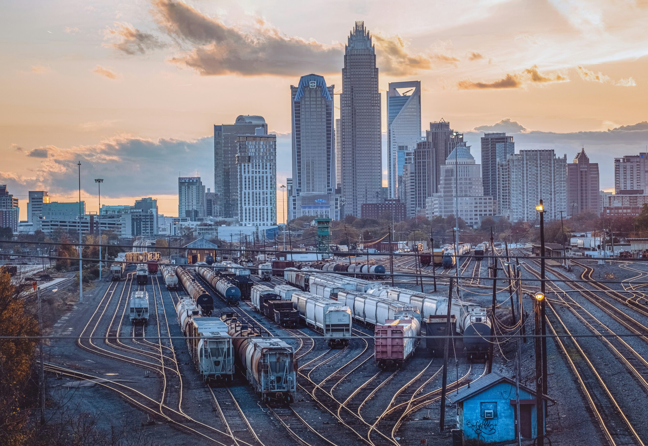 Train cars on tracks in a rail yard with a city skyline and tall buildings in the background at sunset.