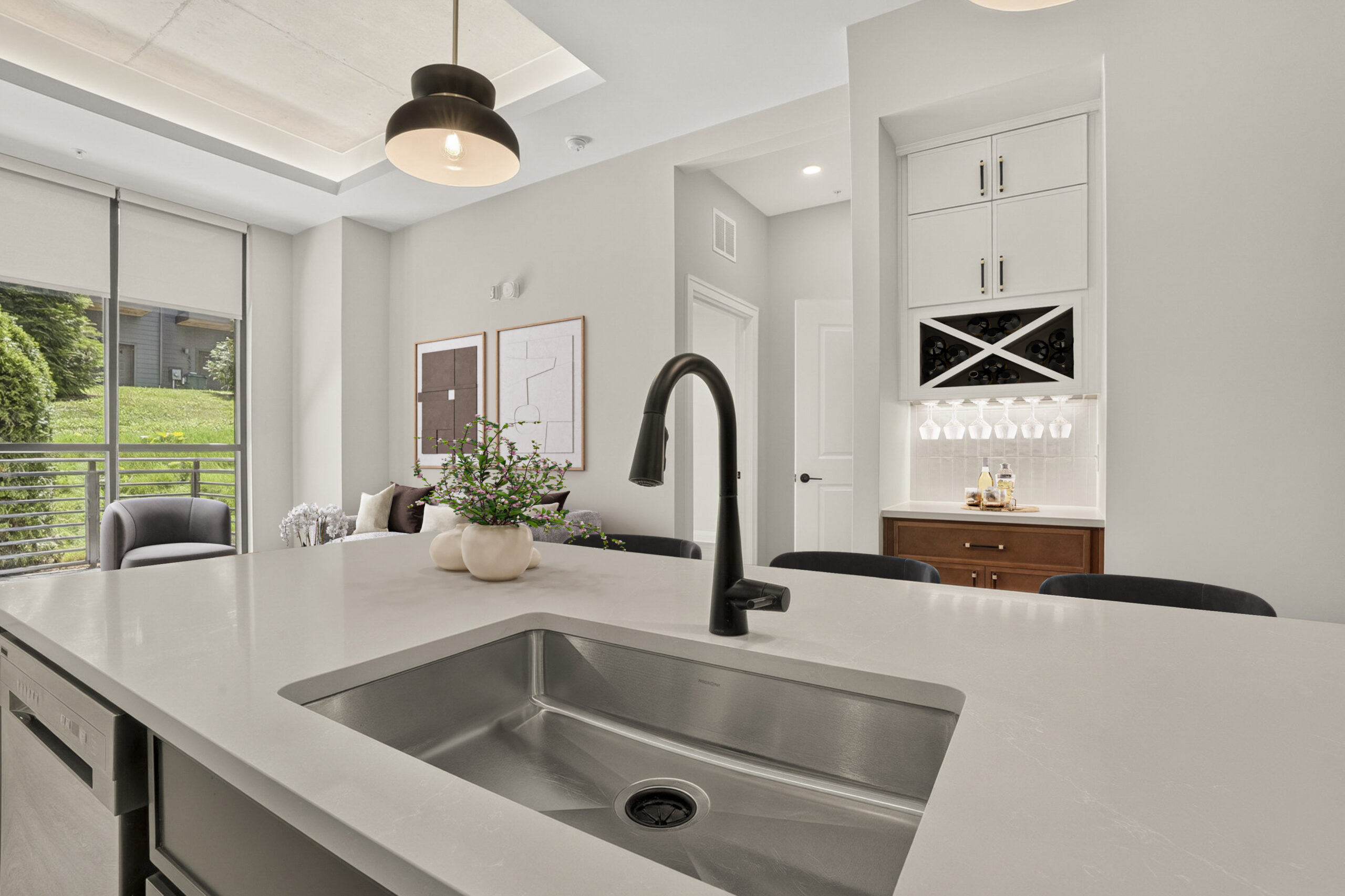 Modern kitchen with a large island sink, black faucet, bar area, pendant lights, and bright windows overlooking greenery.