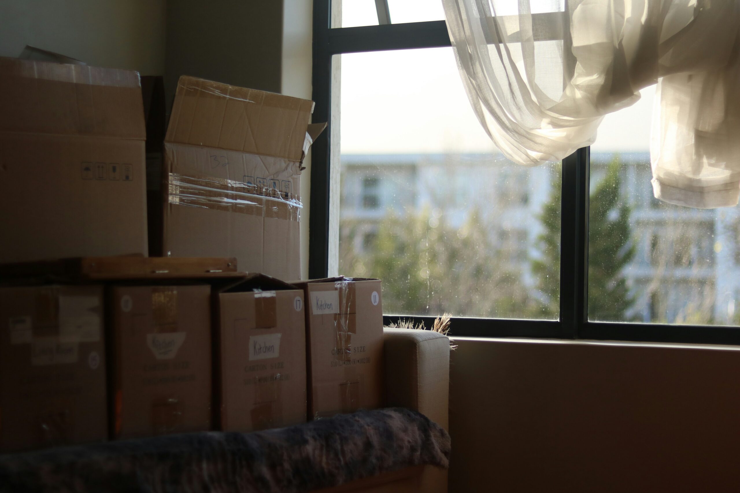 Moving boxes stacked near a window with sheer curtains blowing in sunlight.