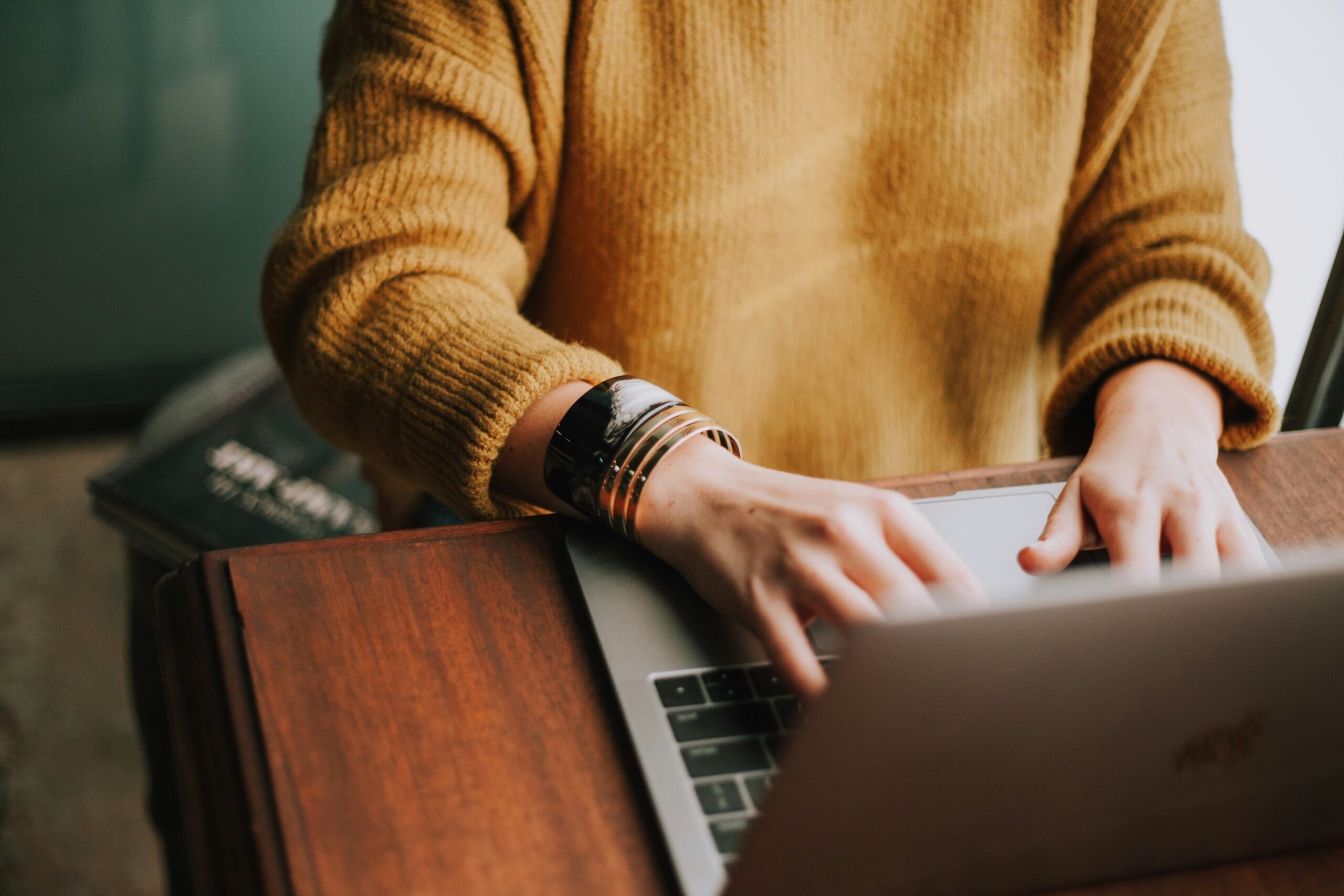 A person in a yellow sweater types on a laptop at a wooden desk, with a book partially visible nearby.