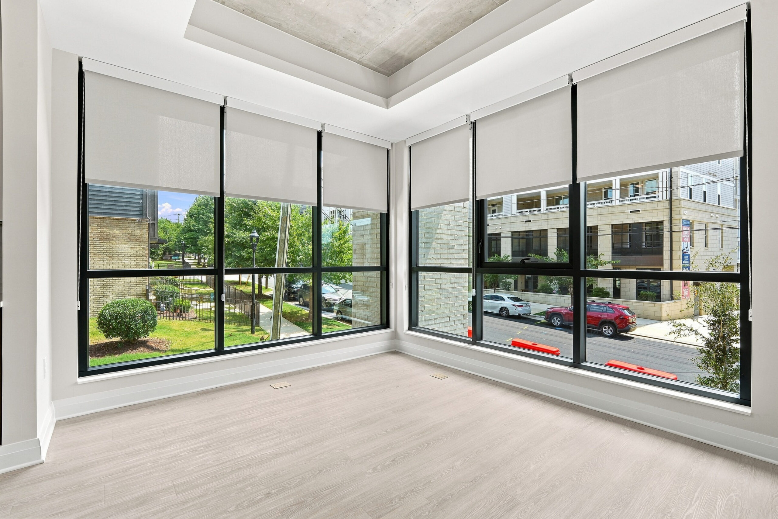 Bright, empty corner room with large floor-to-ceiling windows, light wood flooring, and views of trees and cars outside.