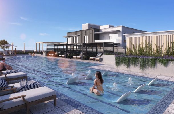 People relax on lounge chairs and in a modern rooftop pool with fountains, under a clear blue sky.