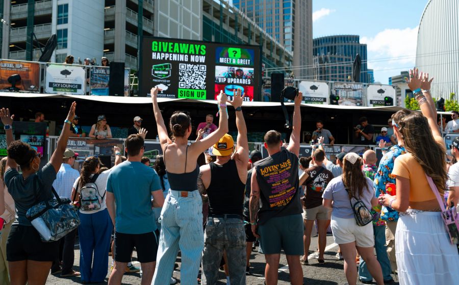A crowd of people facing a large digital screen with event information and a QR code at an outdoor festival.