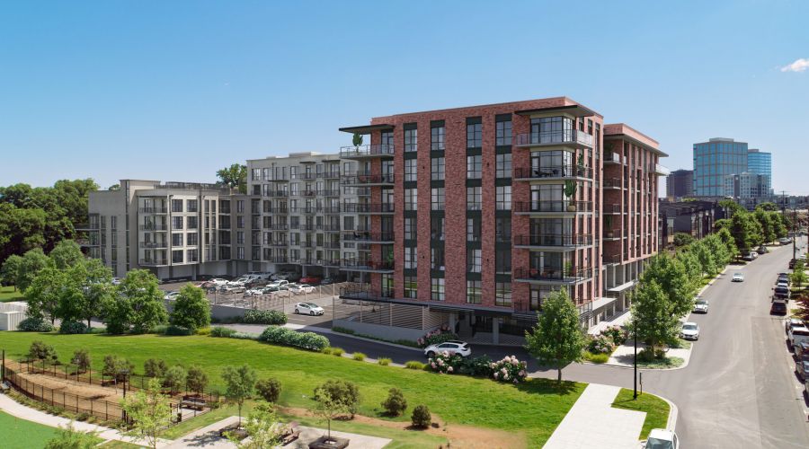 Modern apartment buildings with balconies overlook a green park and street lined with trees and parked cars on a sunny day.
