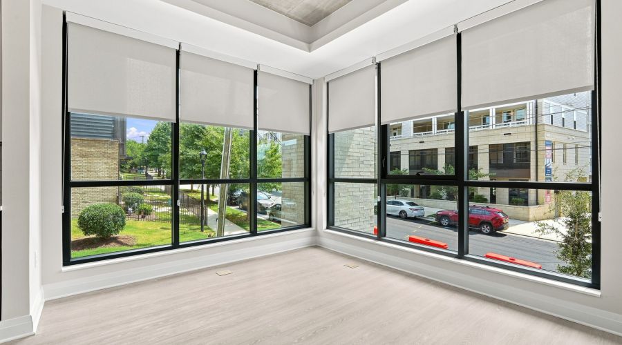 Bright, empty corner room with large floor-to-ceiling windows, light wood flooring, and views of trees and cars outside.