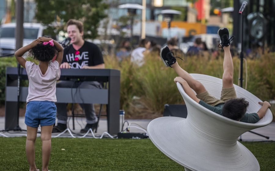 A child in a pink shirt watches a man playing a keyboard while another child reclines in a white spinning chair.