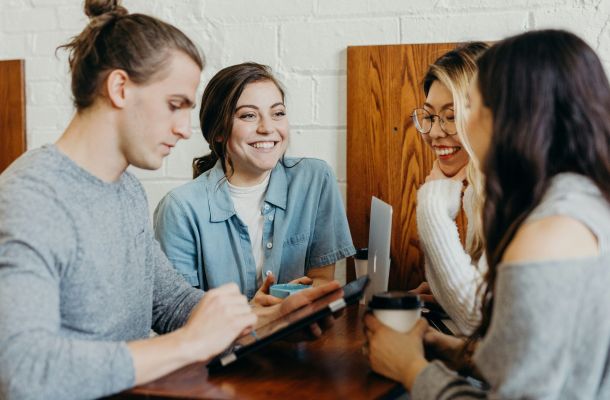 Four young adults sit together at a table, chatting and laughing, with one person holding a tablet and others holding drinks.