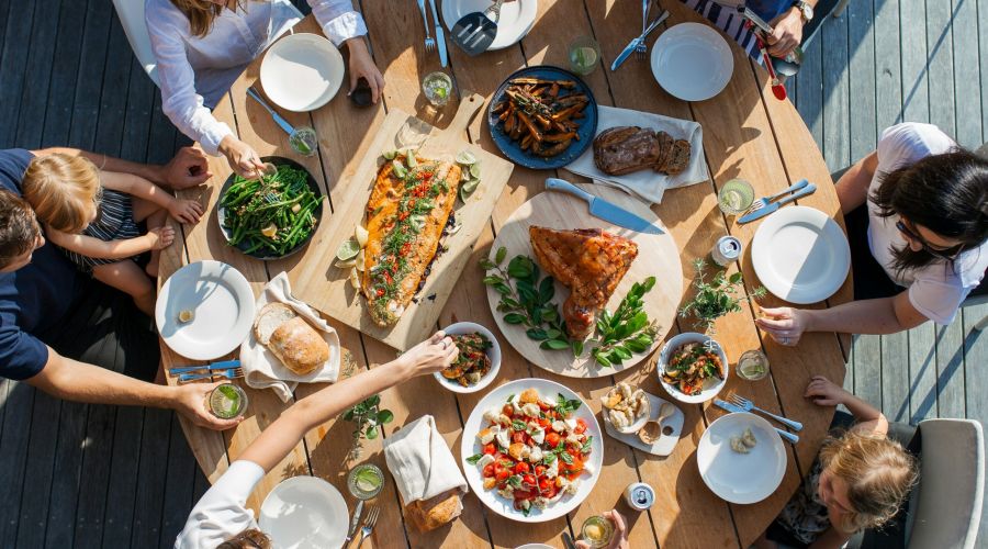 People gathered around a wooden table outdoors, sharing a meal with salads, bread, fish, and drinks.
