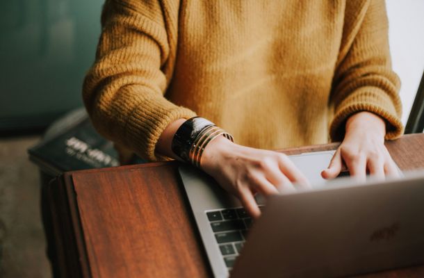 A person in a yellow sweater types on a laptop at a wooden desk, with a book partially visible nearby.