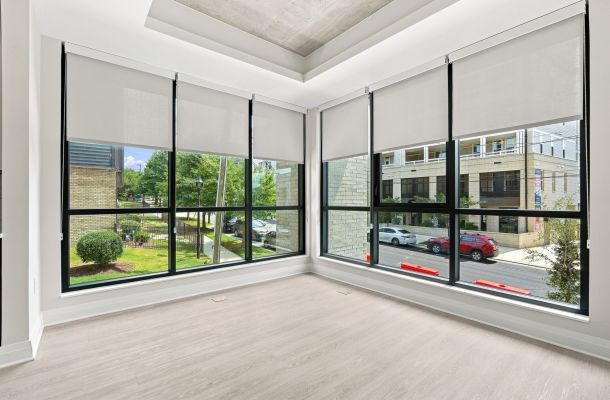 Bright, empty corner room with large floor-to-ceiling windows, light wood flooring, and views of trees and cars outside.
