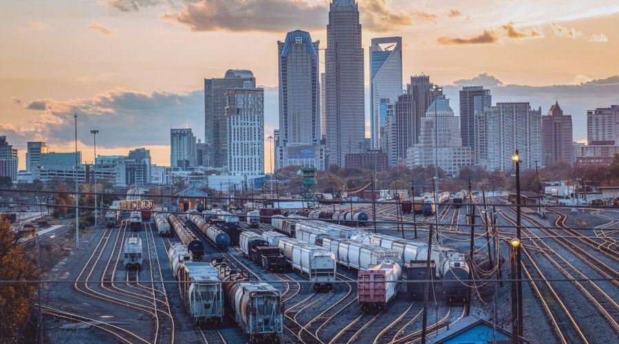 Train cars on tracks in a rail yard with a city skyline and tall buildings in the background at sunset.