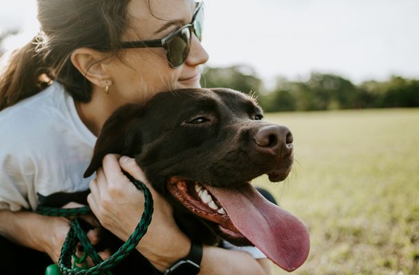 Woman wearing sunglasses hugs a happy brown dog with its tongue out in a grassy field.