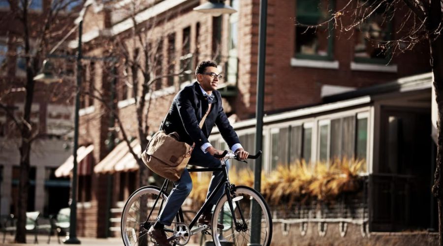 A man in business attire rides a bicycle with a messenger bag in an urban area on a sunny day.