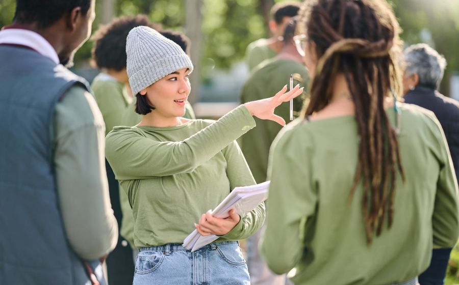 A woman in a grey beanie holds a notebook and gestures while talking to a group of people outdoors.