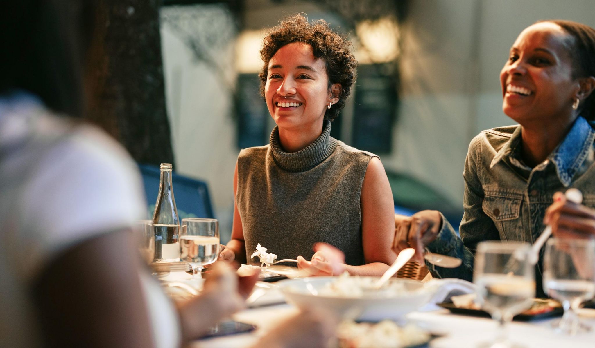 Two women are smiling and enjoying a meal together at a restaurant table with water glasses and plates of food.