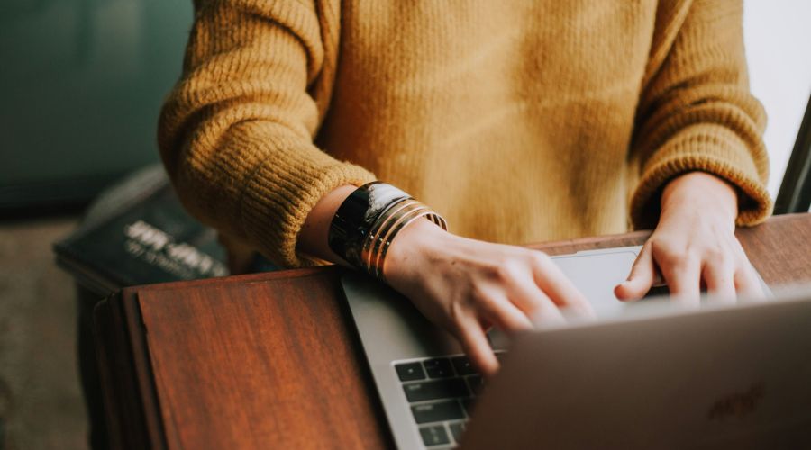 A person in a yellow sweater types on a laptop at a wooden desk, with a book partially visible nearby.